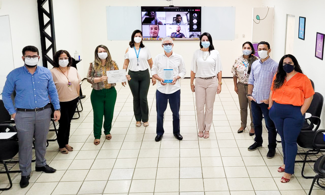 Nove pessoas em pé. Uma mulher segura o certificado. Um homem uma placa. Entrega da política de Diversidade e Inclusão do Inec.