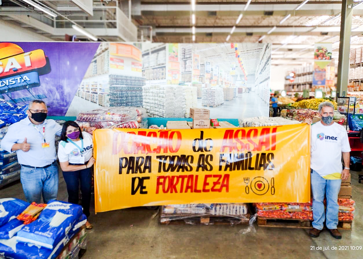 Três pessoas seguram um cartaz com os dizeres: doação do assaí para todas as famílias de fortaleza. Ao fundo, quilos de alimentos não-perecíveis para a Campanha Brasil sem Fome.