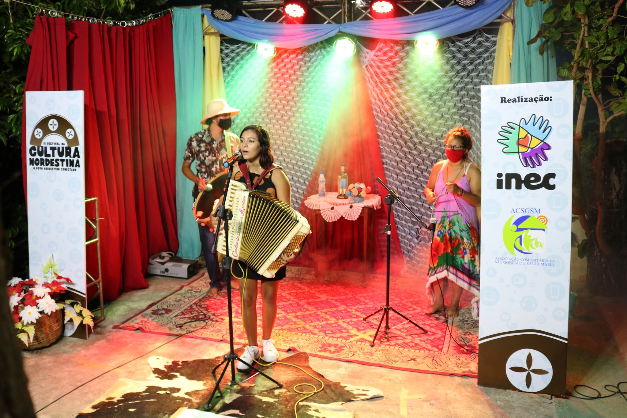 Menina tocando acordeon e cantando durante festival de cultura. Ai fundo, duas pessoas acompanham com instrumentos de percussão como triângulo e zabumba.