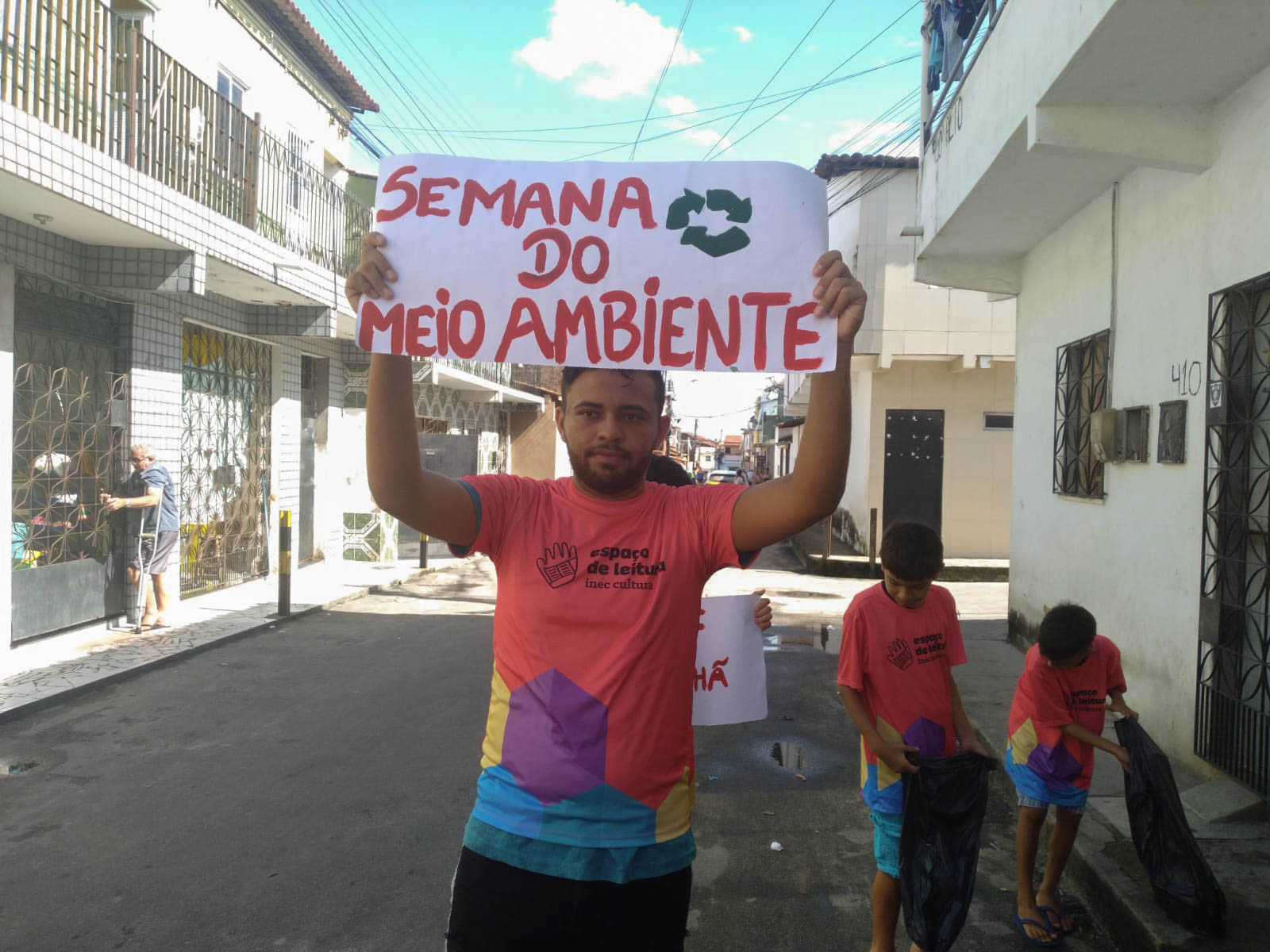 Participantes do Espaço de Leitura Ballet Raio de Sol realizam mutirão d elimpeza no bairro. N imagem, um garoto segura um cartaz escrito Semana do Meio Ambiente, atrás dele dois garotos recolhem lixo na rua