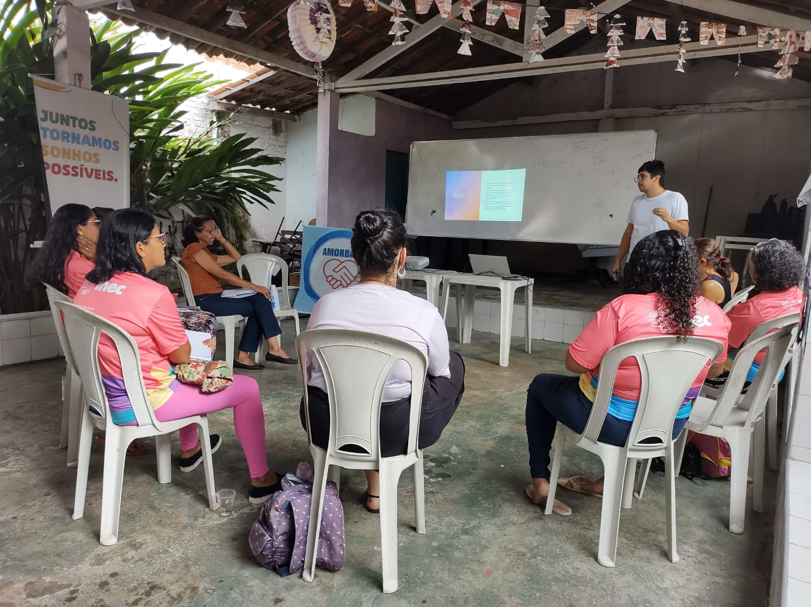 A imagem mostra algumas mulheres sentadas em semi-circulo de frente para uma apresentação em powerpoint