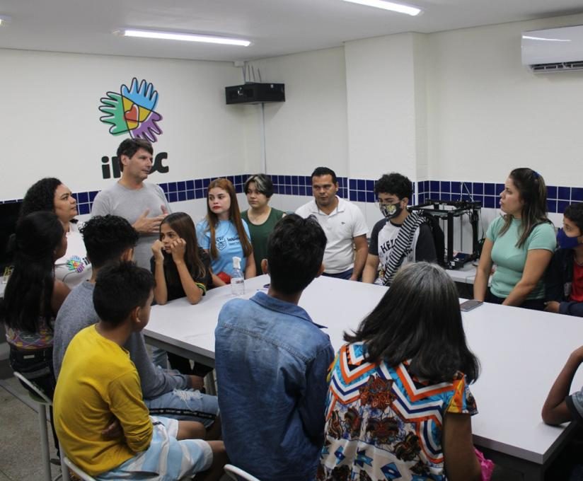 na foto vemos várias familias sentadas ao redor de uma mesa retangular branca em uma sala branca com o símbolo do inec atrás