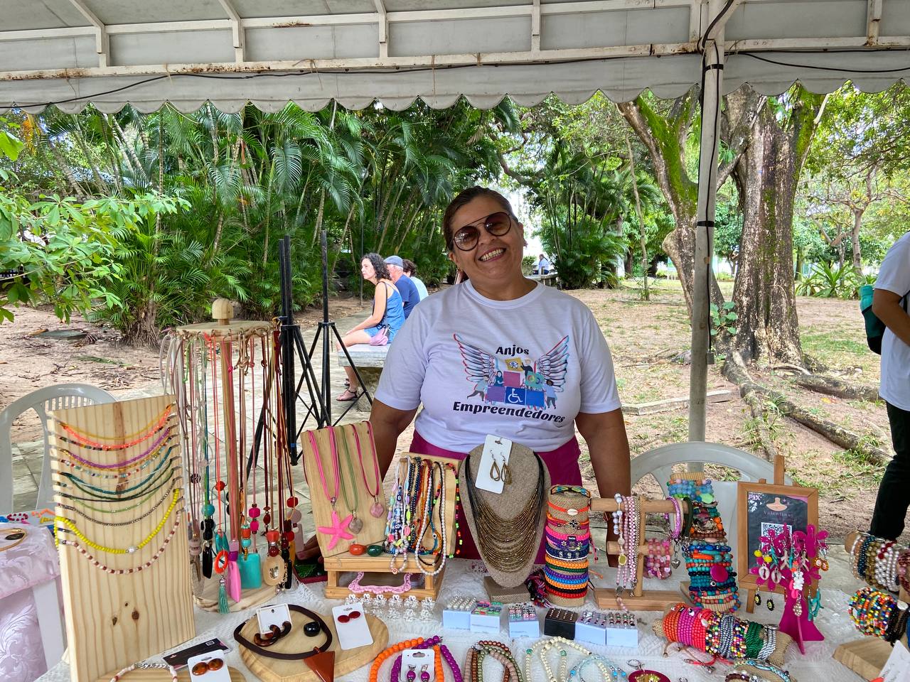 Uma mulher, utilizando uma blusa branca e óculos escuros. Ela está vendendo produtos de miçanga na Feira do Zé