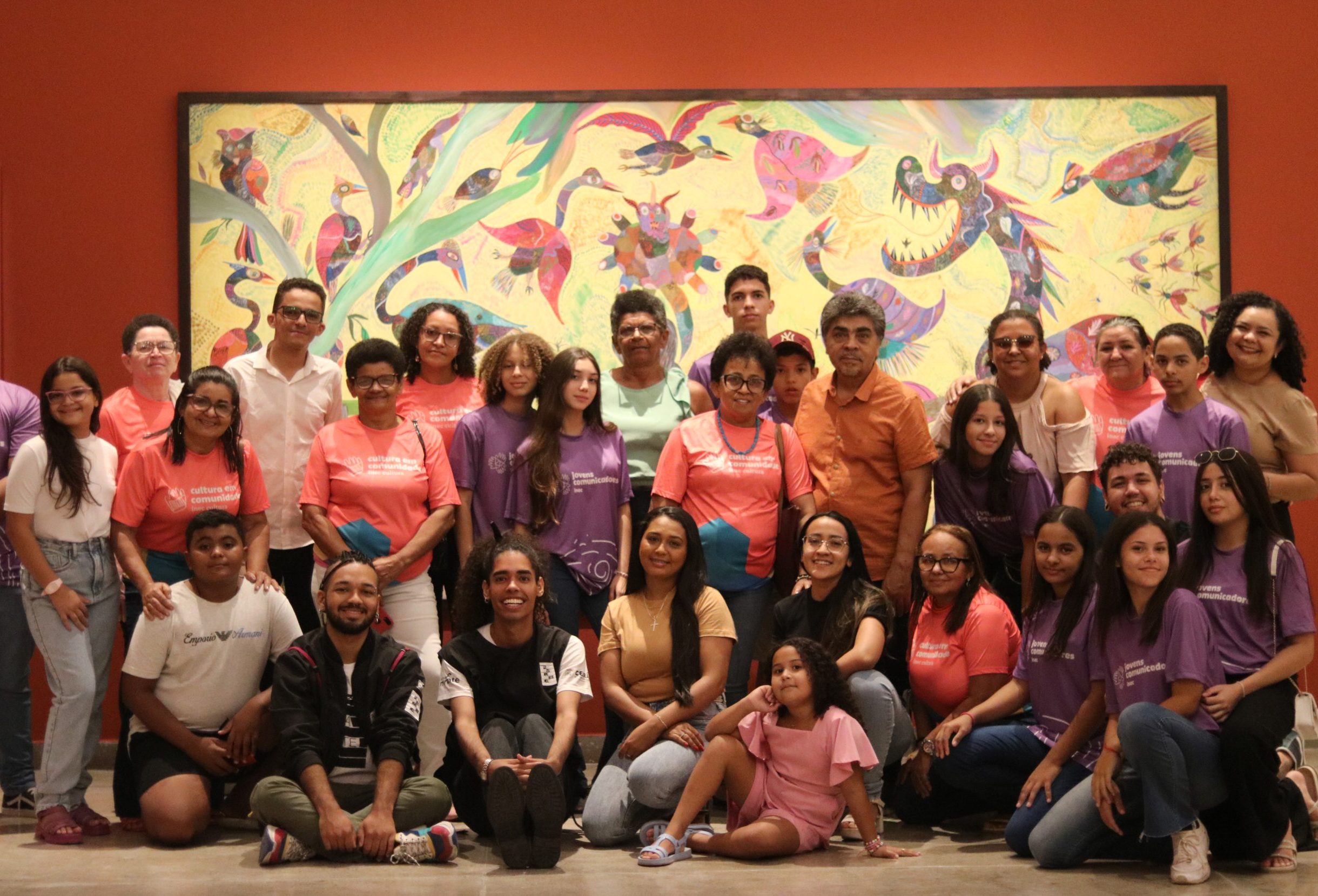 A foto mostra uma turma mista de beneficiários do cultura em comunidade e jovens comunicadores durante visita a equipamentos culturais. A turma posa em frente ao quadro de Chico da Silva