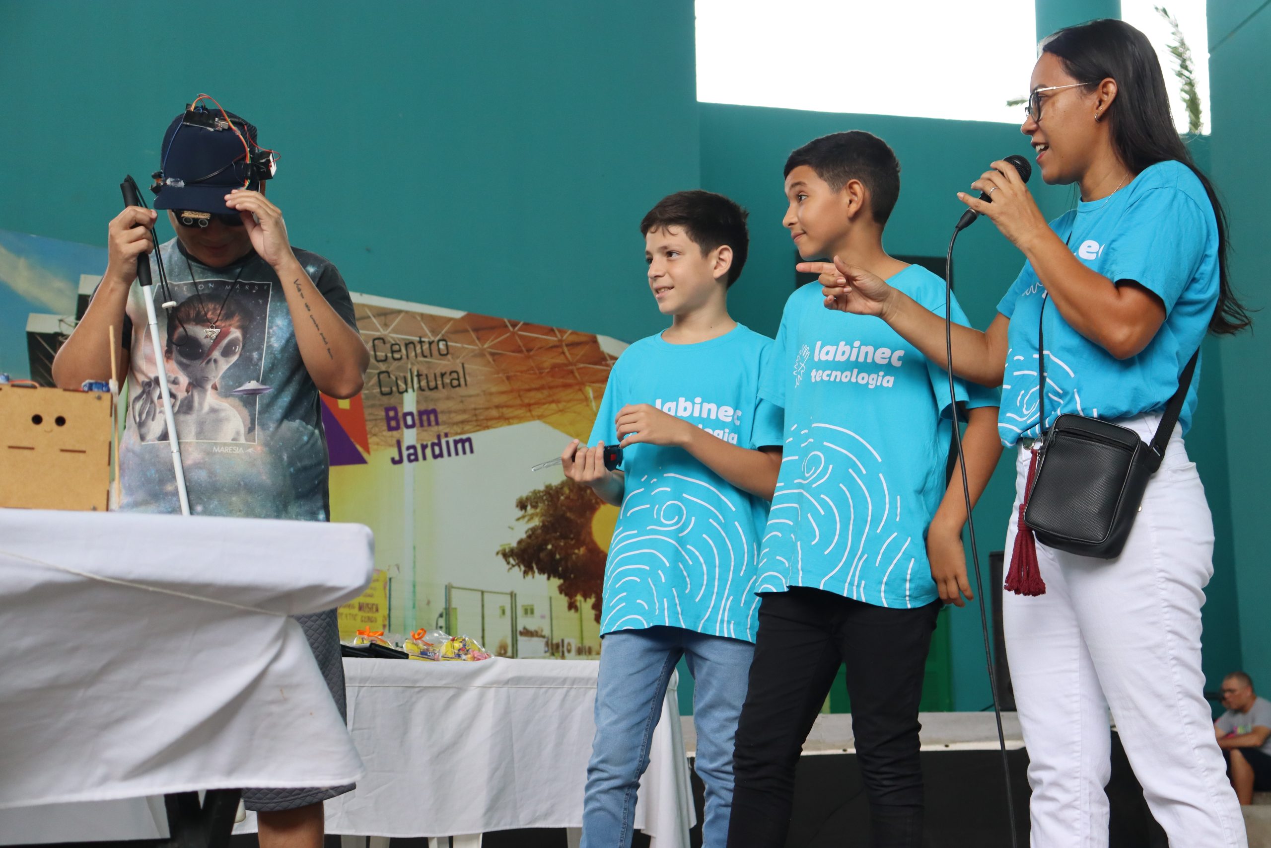 alunos do Labinec em pé ao lado de um deficiente visual que veste o boné do projeto de acessibilidade criado pelos alunos. Ao lado dos dois meninos, Josilda está com um microfone na mão. Todos estão com uma camisa azul escrito Labinec